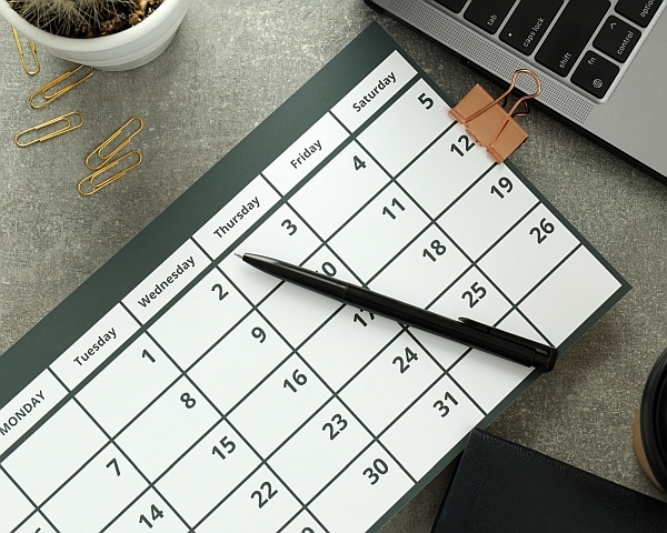 A calendar sitting on a desktop with a pen on top, as if someone is planning a timeline. There is a coffee cup and a laptop nearby